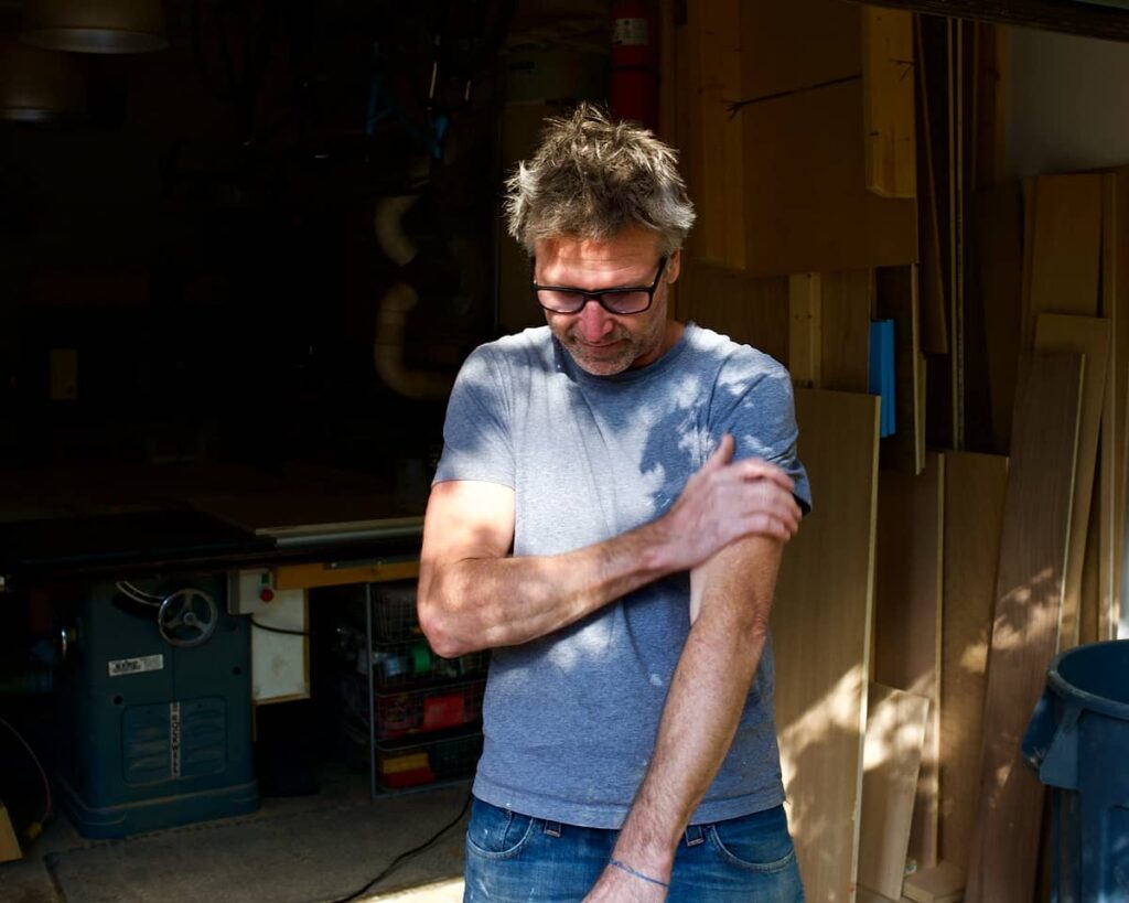 Man in grey t-shirt rubs his arms in front of his workshop