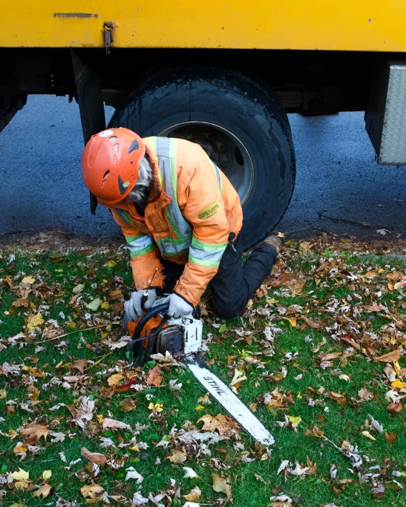 Arborist in an orange coat kneels in the grass and fixes a chainsaw