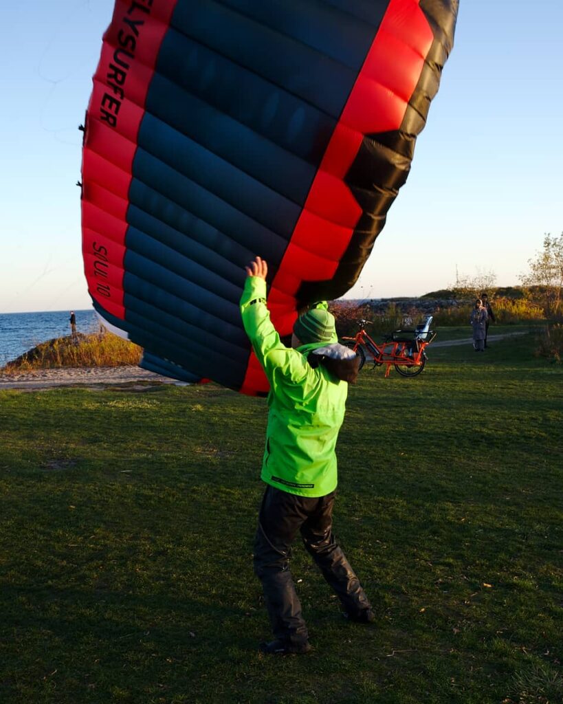Man in black pants and a green coat catches a blue and red kiteboarding kite before if falls to the ground